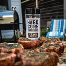 A bottle of Hardcore Carnivore Camo Game & Lamb Seasoning sits outdoors on a table with raw, herbed steaks nearby. A grill, chair, and the exterior of a house appear in the background.