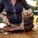 Wearing a dark patterned shirt, a person slices juicy grilled meat seasoned with Hardcore Carnivore Camo Game & Lamb Seasoning by Hardcore Carnivore on a wooden board outdoors.
