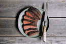 A plate of medium-rare steak with a browned crust, seasoned with Hardcore Carnivore Camo Game & Lamb Seasoning, sits beside a rustic knife on a green plate atop a weathered wooden surface.