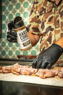 A person in black gloves and a camouflage shirt sprinkles Hardcore Carnivore Camo Game & Lamb Seasoning by Hardcore Carnivore onto raw meat on a countertop, with a decorative tile wall in the background.