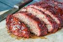 A juicy, freshly cooked meatloaf seasoned with Hardcore Carnivore Red All Purpose Seasoning is sliced on a wooden cutting board beside a large knife, its shiny glazed topping making it look irresistible.