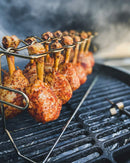 Chicken drumsticks, seasoned with Hardcore Carnivore Red All Purpose Seasoning by Hardcore Carnivore, are stacked vertically on a grill rack over a barbecue, with smoke wafting in the background.