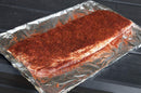 A raw slab of pork ribs coated in Hardcore Carnivore Red All Purpose Seasoning sits on aluminum foil, ready to be cooked, on a dark table.