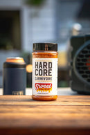 A bottle of Hardcore Carnivore Sweet BBQ Rub by Hardcore Carnivore rests on a wooden outdoor surface, promising bold barbecue flavor. A blurred drink container and grill are visible in the background.