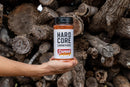 A hand holds a container of Hardcore Carnivore Sweet BBQ Rub, featuring a black lid and a white label with bold black and red text, in front of stacked firewood. This flavorful BBQ seasoning is crafted by Hardcore Carnivore.