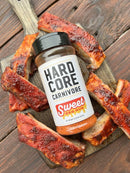 A bottle of Hardcore Carnivore Sweet BBQ Rub by Hardcore Carnivore is placed on a wooden board, with glazed pork ribs highlighting its signature barbecue flavor atop a rustic table.
