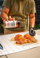 Wearing a green shirt and a black glove, someone sprinkles Hardcore Carnivore Sweet BBQ Rub by Hardcore Carnivore onto raw chicken pieces on a cutting board outdoors.