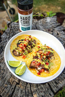 A white plate holds two tacos with grilled meat seasoned with Hardcore Carnivore Tex-Mex Taco and Fajita Seasoning, diced vegetables, sauce, and lime wedges. In the background, a Hardcore Carnivore container rests on a tree stump.