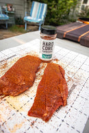 Two raw steaks coated with Hardcore Carnivore Tex-Mex Taco and Fajita Seasoning sit on a cutting board beside a jar of the same seasoning by Hardcore Carnivore, with outdoor furniture and greenery in the background.