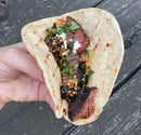 A hand holds a grilled steak taco in a soft flour tortilla, topped with chopped onions, cilantro, and Hardcore Carnivore Tex-Mex Taco and Fajita Seasoning by Hardcore Carnivore, against a weathered wooden background.