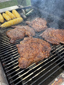 Four steaks seasoned with Hardcore Carnivore Tex-Mex Taco and Fajita Seasoning grill alongside corn and a green chili pepper, as smoke rises from the barbecue.