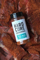 A bottle of Hardcore Carnivore Tex-Mex Taco and Fajita Seasoning rests atop raw meat, which is generously coated with the same reddish seasoning. The label clearly shows the Hardcore Carnivore brand.
