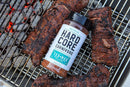 A bottle of Hardcore Carnivore Tex-Mex Taco and Fajita Seasoning rests on a grill beside charred steaks and glowing coals—ideal for bringing bold Tex-Mex flavor to your next cookout.