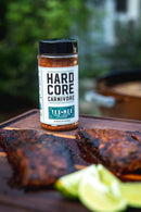 A bottle of Hardcore Carnivore Tex-Mex Taco and Fajita Seasoning by Hardcore Carnivore sits on a wooden surface next to grilled meat and lime wedges, ready to add bold taco flavor to your favorite dishes.