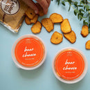 A hand grabs a toasted bread slice beside two In The Curious Kitchen Beer Cheese containers and a bag of artisan pretzels, all from the In the Curious Kitchen Beer Cheese & Pretzels Bundle, set against an orange backdrop with a green plant.
