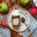 A jar of In The Curious Kitchen Bourbon Apple Butter sits on a decorative plate with a spoonful beside it, surrounded by green apples and a colorful floral cloth—ideal for charcuterie boards and Southern entertaining.