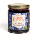 A glass jar of In The Curious Kitchen Bourbon Blueberry Jam features a gold lid and a label with blueberries, the product name, and net weight 9 oz (255g), highlighting its rich Kentucky bourbon flavor.