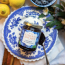 A jar of In The Curious Kitchen Bourbon Blueberry Jam sits on a blue and white plate with a spoon, surrounded by lemons, blueberries, and flowers. This delicious jam is infused with Kentucky bourbon—perfect for your table.