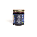 A glass jar of In the Curious Kitchen Bourbon Blueberry Jam is shown. The label features "Jam Packed With Flavor," blue and green designs, and displays the website and social media info at the bottom.