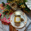 A jar of In The Curious Kitchen Bourbon Honey Mustard sits on a decorative plate with a spoon beside it. An open jar, yellow flowers, and colorful cloths are arranged nearby on a wooden and marble surface.