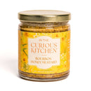 A glass jar with a gold lid holds In The Curious Kitchen Bourbon Honey Mustard. Its yellow, floral label shows a net weight of 9 oz (255 g). Handcrafted in Kentucky, it’s perfect for adding to your charcuterie board.