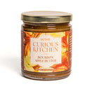 A glass jar with a gold lid, part of the In The Curious Kitchen Bourbon-Infused Gourmet Gift Set, features apples and Kentucky bourbon. This 9 oz treat has an orange and cream label and makes a unique gourmet gift.