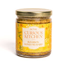 A glass jar of In The Curious Kitchen Bourbon Honey Mustard (9 oz/255g) with floral yellow label and gold lid—an ideal addition to the In the Curious Kitchen Bourbon-Infused Gourmet Gift Set.