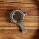 The In the Curious Kitchen Cocktail Bar Tool Kit by In The Curious Kitchen features a stainless steel cocktail strainer with a spring coil, shown on a striped wooden surface—an essential for any home bartender.