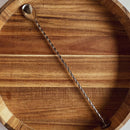The In the Curious Kitchen Cocktail Bar Tool Kit by In The Curious Kitchen features a long, twisted metal bar spoon, shown here resting diagonally in an empty wooden bowl—an essential for home bartenders.