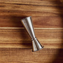 The In The Curious Kitchen Cocktail Bar Tool Kit features a metal jigger on a wooden surface with visible grain—ideal for home bartenders and mixology fans.