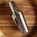 The In the Curious Kitchen Cocktail Bar Tool Kit by In The Curious Kitchen features a shiny stainless steel cocktail shaker on a round wooden tray showcasing natural wood grain patterns.