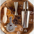 The In the Curious Kitchen Cocktail Bar Tool Kit by In The Curious Kitchen includes a wooden tray with cocktail shaker, jigger, citrus reamer, strainer, mixing spoon, dried citrus slices, fresh cherries, and ice cubes—ideal for home bartenders.