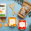 A flat lay on a blue background features two wooden spoons, crackers, pretzel crisps, green leaves, and the In The Curious Kitchen Derby Party Bundle 2-Pack—two tubs of cheese spreads ideal for charcuterie boards or Southern gifting.