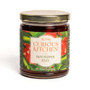 A 9 oz jar of In The Curious Kitchen Hot Pepper Jelly, handcrafted in Kentucky in small batches, with a gold lid and a label showing green peppers on a red background.