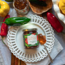 A jar of In The Curious Kitchen Hot Pepper Jelly sits on a white decorative plate with green and red peppers, a spoonful of jelly, and another open jar, all arranged on a wooden table.