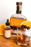 A bottle of Woodford Reserve whiskey, In the Curious Kitchen Orange Bitters by In The Curious Kitchen, and an old fashioned cocktail with orange slices and cherries—everything you need for classic bourbon cocktails at home.