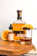 A bottle of In The Curious Kitchen Orange Bitters sits beside an old fashioned cocktail garnished with orange and cherries—perfect inspiration for home bartending and bourbon cocktails.