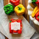 A 2-pack of In The Curious Kitchen Pimento Cheese rests on parchment beside fresh green, yellow, and red bell peppers on a wooden surface with a striped cloth—ideal for a classic Southern snack.