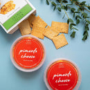 The In The Curious Kitchen Pimento Cheese & Crackers Bundle features two tubs of pimento cheese and Rustic Bakery crackers on a light blue surface with scattered crackers and green leaves, perfect for charcuterie or classic Southern snacking.