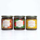 The In The Curious Kitchen Southern Charcuterie Spread Bundle features three colorful jars—Bourbon Apple Butter, Hot Pepper Jelly, and Bourbon Honey Mustard—displayed side by side with green lids against a white background.