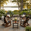 Four Kanyon Living Adirondack Rocking Chairs in Black & Deluxe Antique Mahogany surround a fire pit on the stone patio. Cozy blankets, potted plants on a small table, string lights, and lush blooms complete this lakeside retreat.