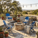 A cozy outdoor patio with Kanyon Living Adirondack Rocking Chairs in Blue & Light Grey around a stone fire pit, potted cacti nearby, string lights above, and scenic hills under a clear sky.