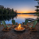 Five Kanyon Living Adirondack Rocking Chairs in Dark Green & Cedar Wood encircle a campfire by a serene lake at sunset, with pine trees, a dock, and canoes in the background. Crafted for durability by Kanyon Living, these seats invite outdoor relaxation.