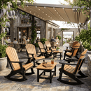 A cozy outdoor patio features Kanyon Living Adirondack Rocking Chair Deluxe in Antique Mahogany & Black. Surrounded by plants, guests dine near a rustic building with large windows beneath a canopy of string lights.