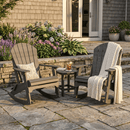 Two Kanyon Living Adirondack Rocking Chair Deluxe Driftwood Grey chairs with cushions and a knit blanket sit on a stone patio by a small round table, surrounded by plants and flowers near a house with open doors.