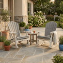 Two Kanyon Living Adirondack Rocking Chair Deluxe Driftwood Grey & White rockers, one with a throw blanket, sit by a small table with a drink and book on a stone patio amid potted plants and flowers near a house.