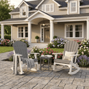 Two Kanyon Living Adirondack Rocking Chair Deluxe Driftwood Grey & White rockers sit with cushions and a blanket on a stone patio by a beige house. Colorful flowers and shrubs surround the space, while a small table between the chairs holds two cups.