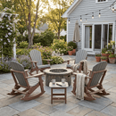 Four Kanyon Living Adirondack Rocking Chairs in Light Grey & Weathered Wood are arranged around a fire pit on a stone patio with string lights, potted plants, and a small table with mugs and a book. A house with French doors is in the background.