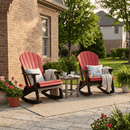 Two Kanyon Living Adirondack Rocking Chairs in red and black sit on a patio by a brick house, with cushions, blankets, and a small table holding mugs and books, surrounded by greenery and flowers for all-weather comfort.