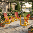 A cozy patio with three Kanyon Living Adirondack Rocking Chairs in red and yellow, a small round table with a drink, a striped blanket on one chair, potted flowers, blooming bushes, and a view of a peaceful lake.